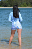 Person wearing a light blue long-sleeve shirt and white floral shorts standing in shallow water near a beach.