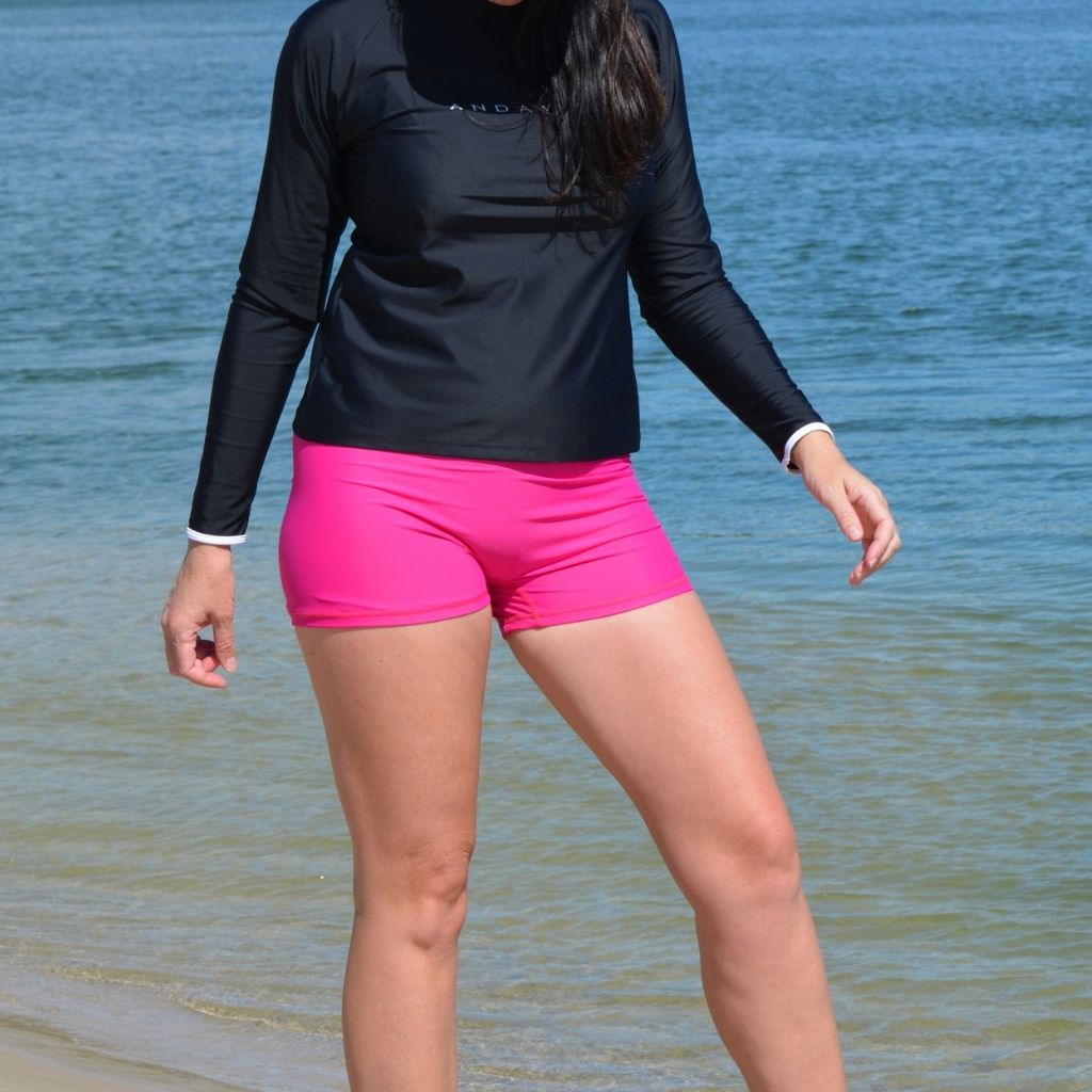 Person wearing a black long-sleeve top and pink shorts standing on a beach.
