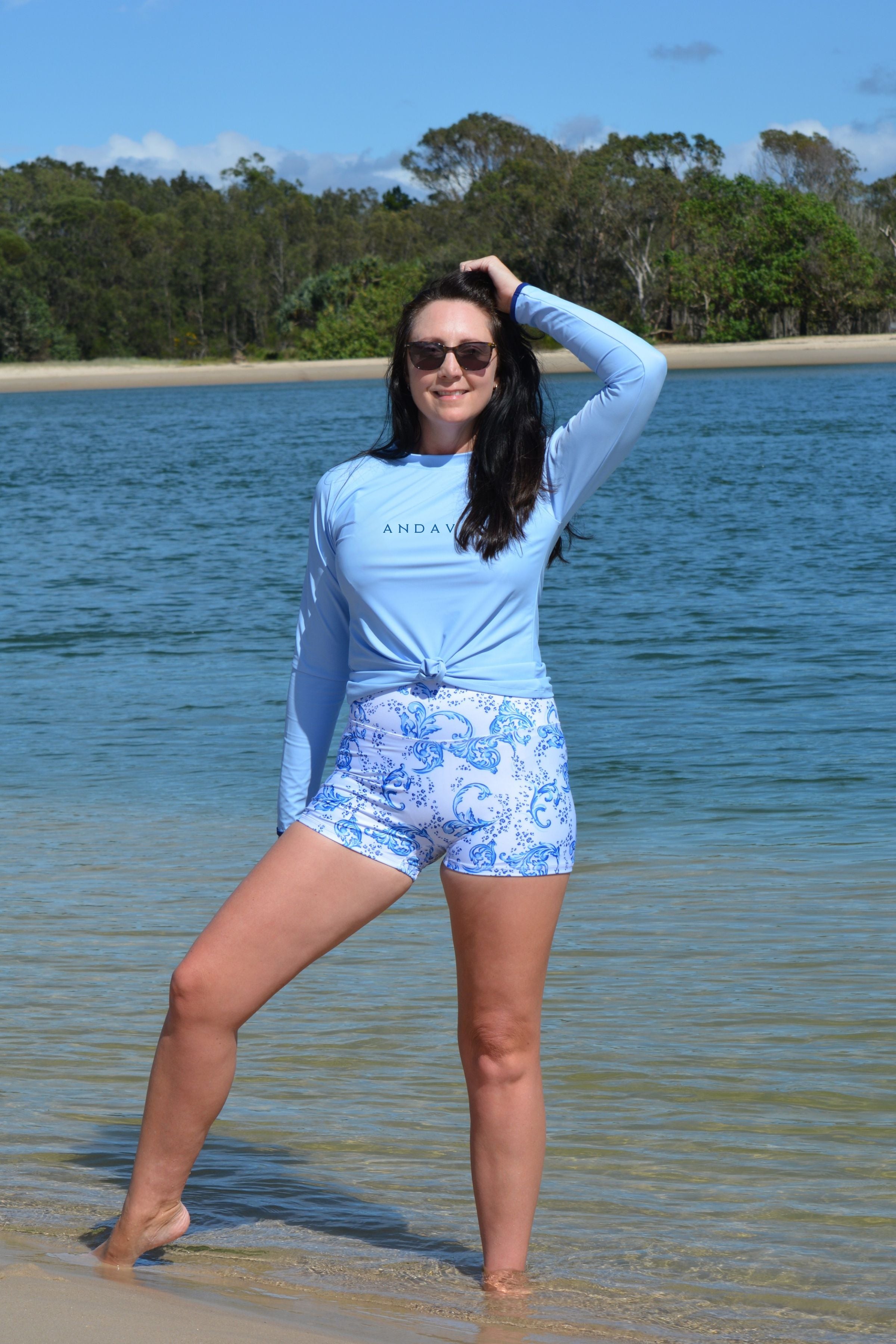 Woman standing on a beach wearing a light blue long-sleeve shirt and floral shorts.