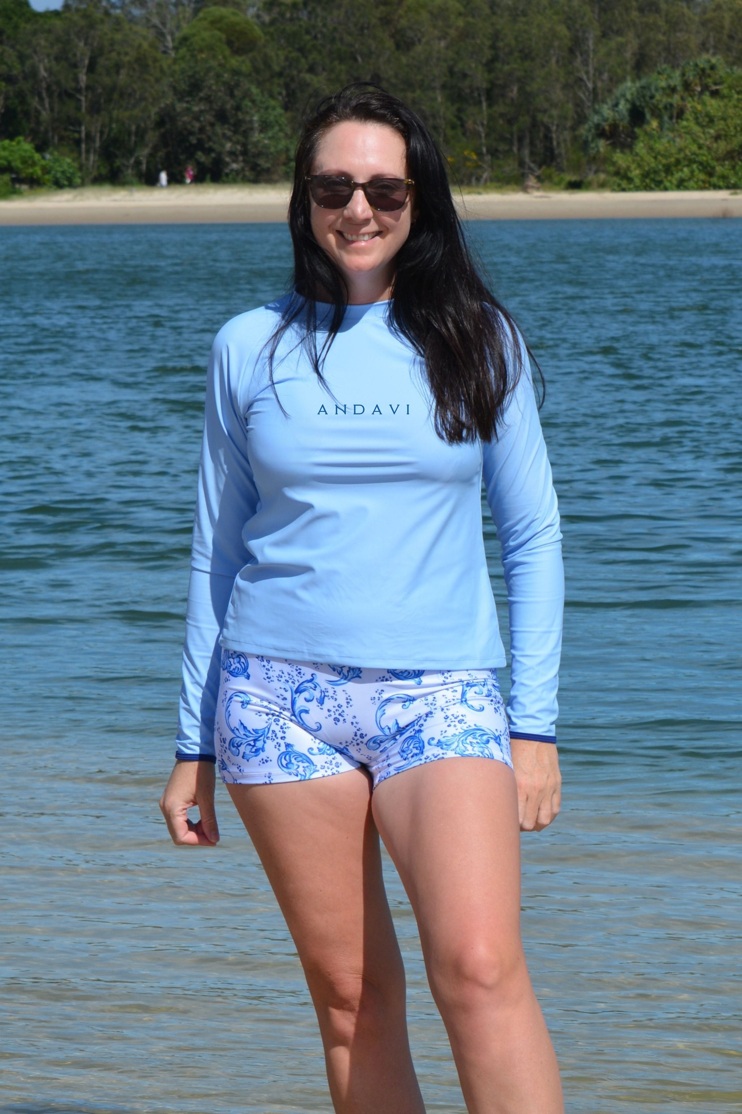 Woman wearing a light blue long-sleeve shirt and patterned shorts standing by a body of water.