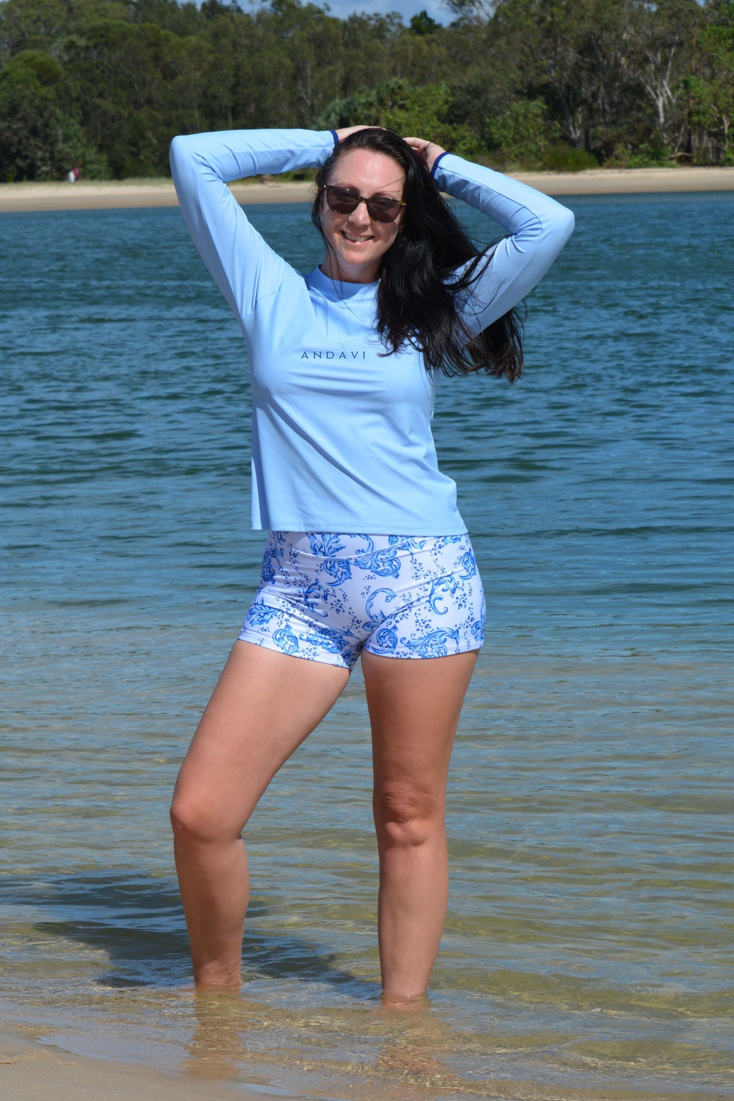 Woman in light blue long-sleeve top and patterned shorts standing in shallow water with trees in the background.