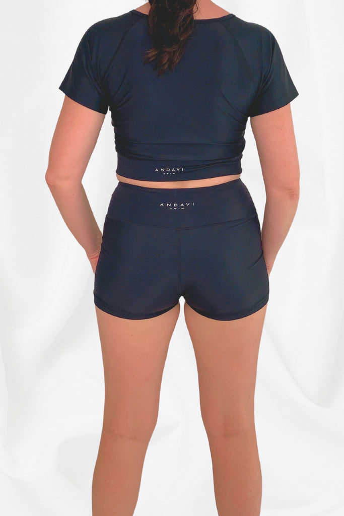 model facing backward wearing Andavi Swim black short sleeve crop top and black swim shorts 