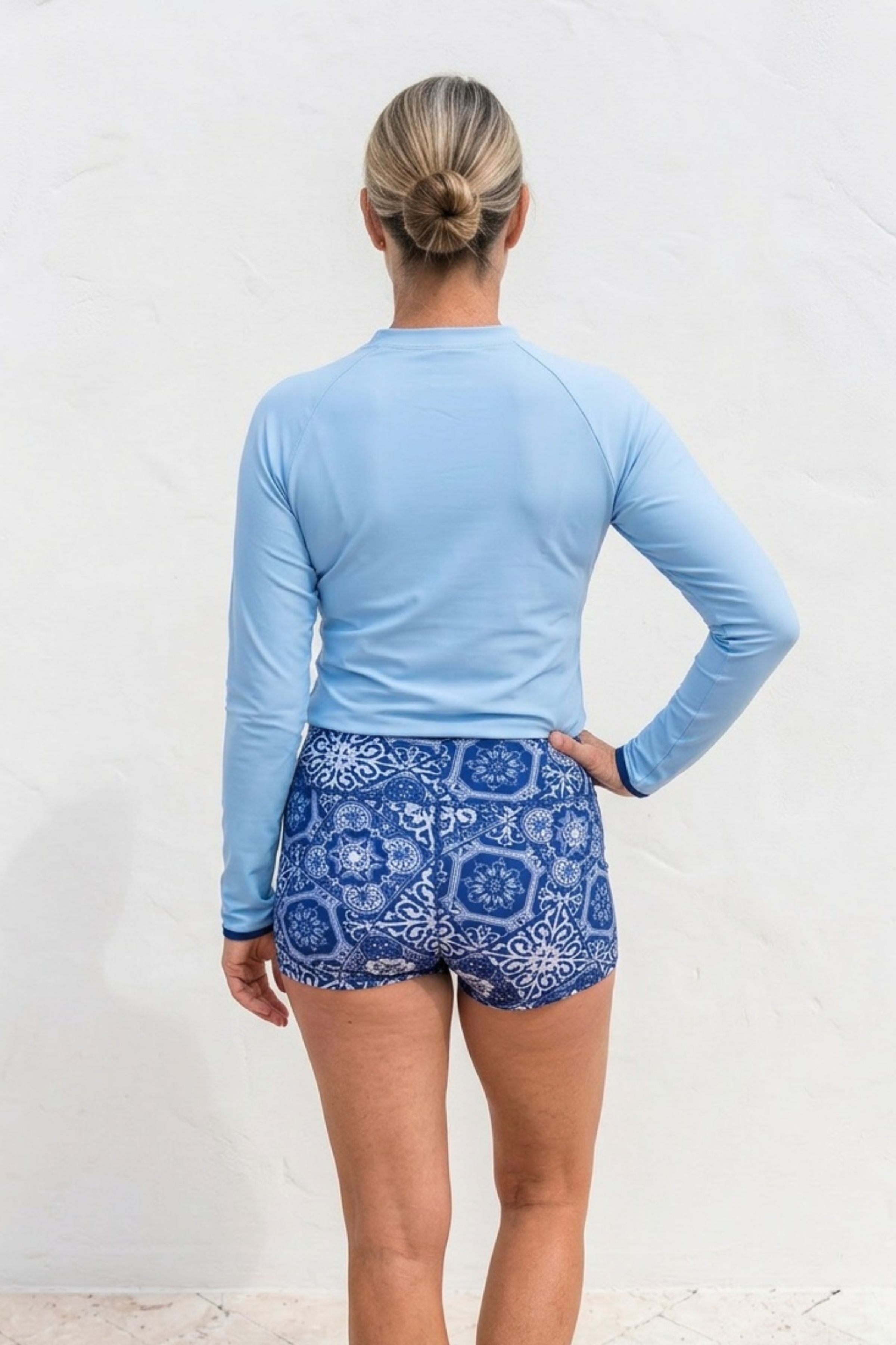 Person wearing a light blue long-sleeve shirt and blue patterned shorts on a white background