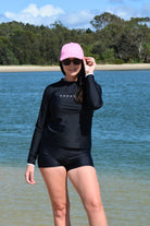 model wearing black andavi swim loose fit rash guard and matching black swim shorts with pink andavi swim logo cap
