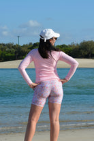 picture of a women standing on the beach with her back to the camera wearing light pink long sleeve rash guard and floral bike shorts