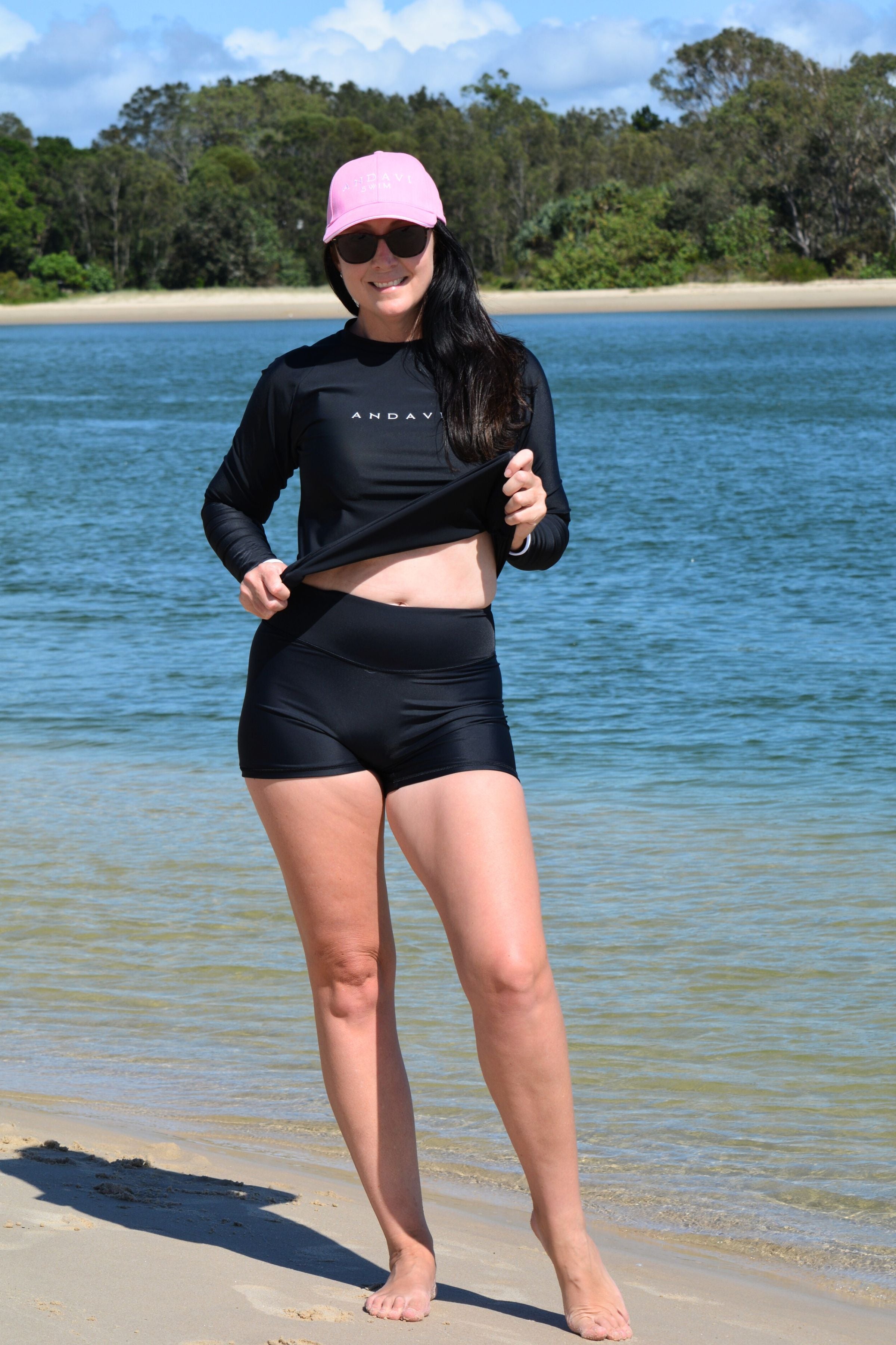 model wearing black andavi swim loose fit rash guard and matching black swim shorts with pink andavi swim logo cap