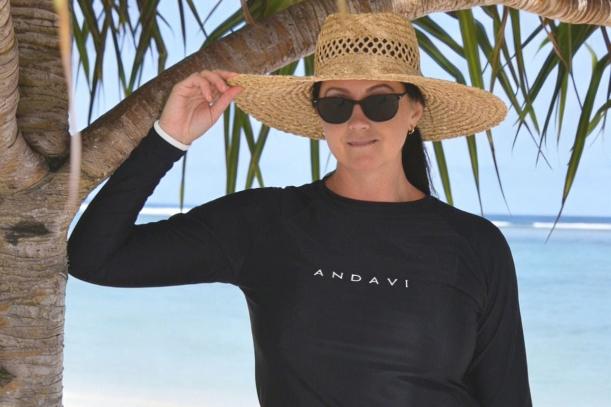 Woman wearing a black long-sleeve rash guard and shorts on a beach with palm trees.