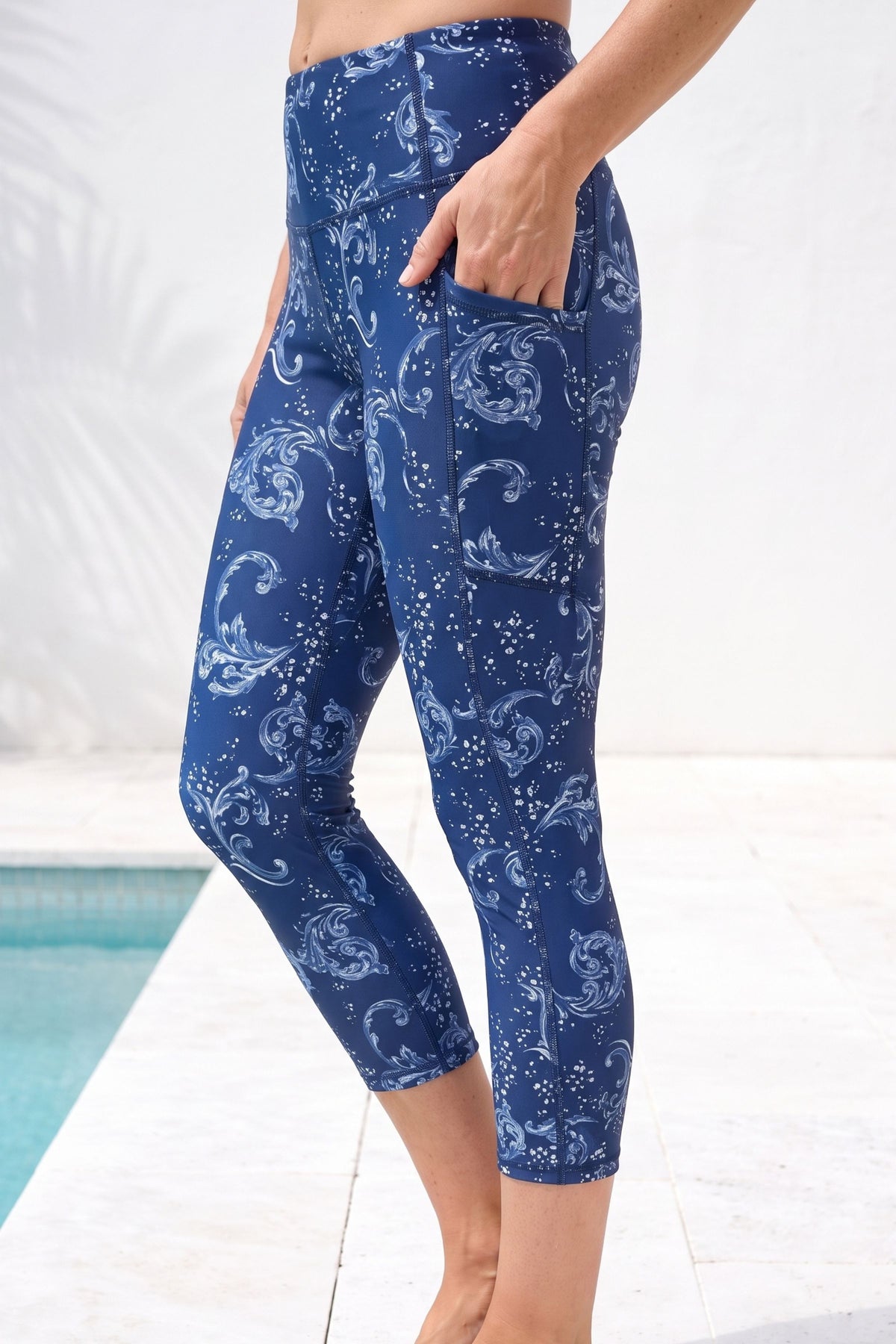 A person wearing blue capri leggings with a white paisley and star pattern, standing by a pool with a white tiled background.