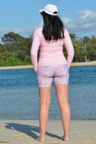 back view of a women standing on the beach wearing light link rash guard and floral bike shorts