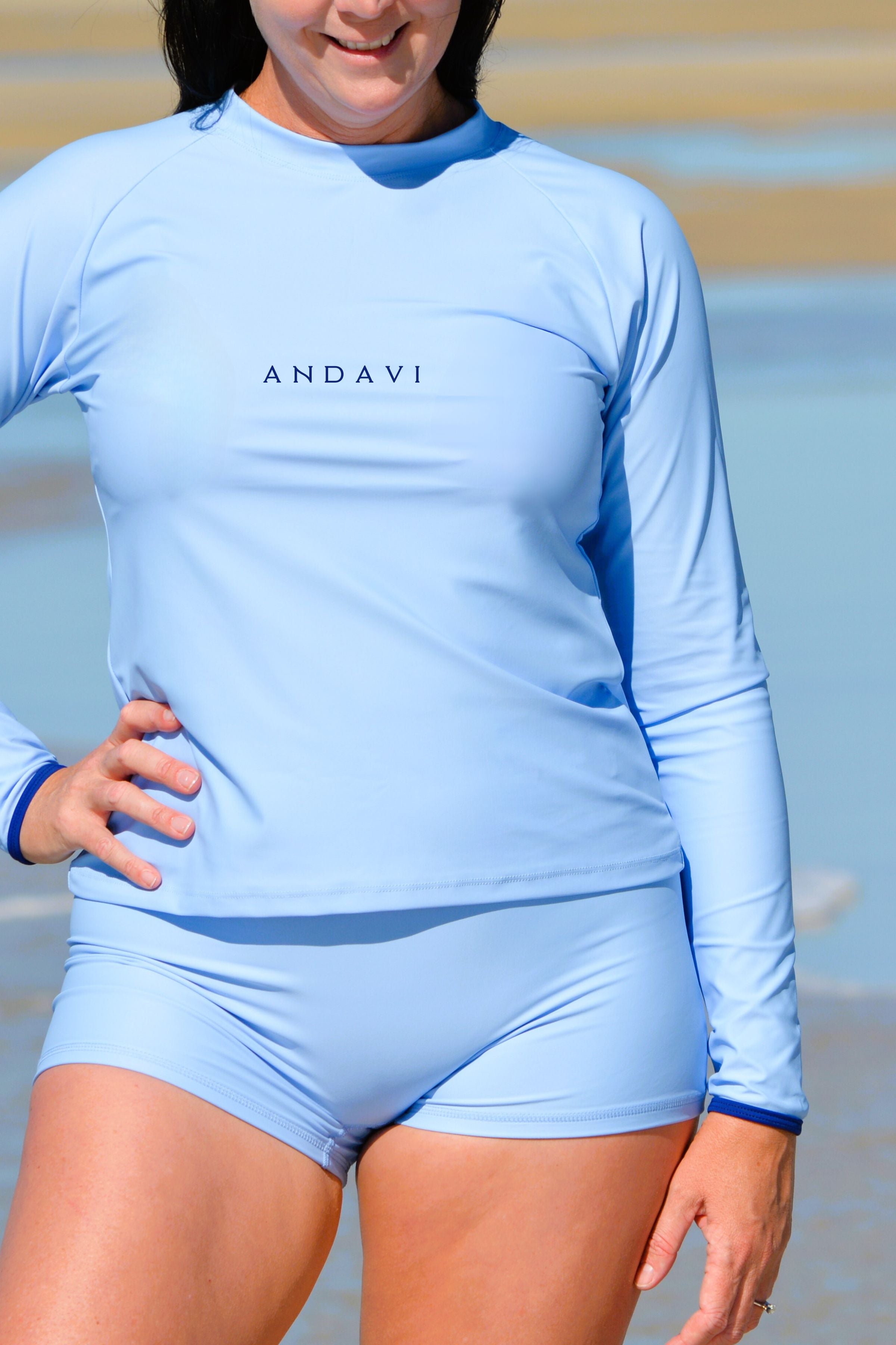 Person wearing a light blue long-sleeve rash guard and shorts with 'ANDAVI' branding on a beach.
