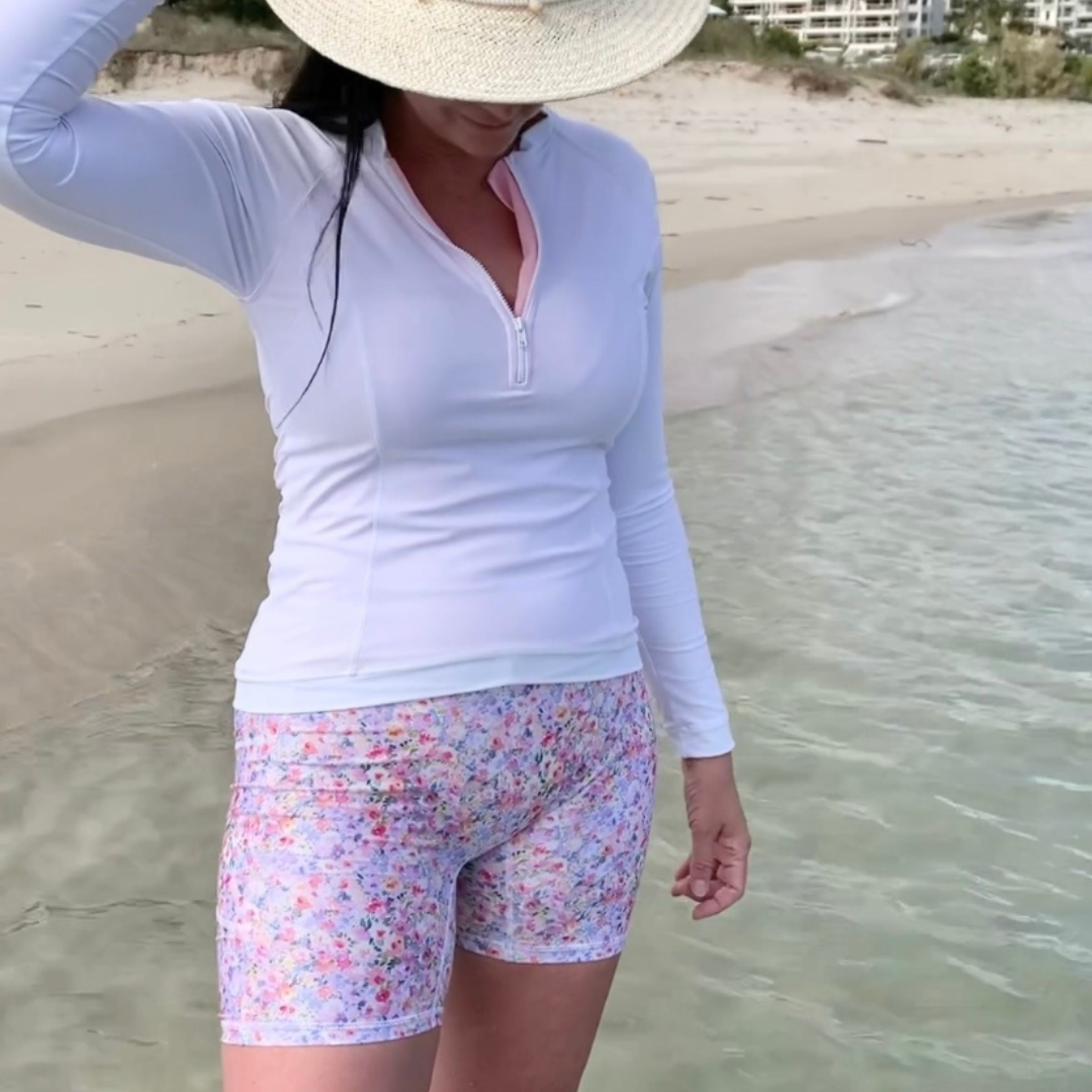 Person wearing a white long-sleeve shirt, floral shorts, and a straw hat on a beach.