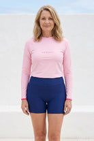 women wearing light pink andavi rashguard with navy blue shorts infront of white textured wall