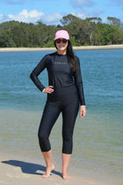 Woman in black capri swim leggings and black andavi rash guard and pink cap standing on a beach with water and trees in the background