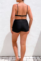 women  wearing black high-waisted swim shorts and a black bikini top against a white background