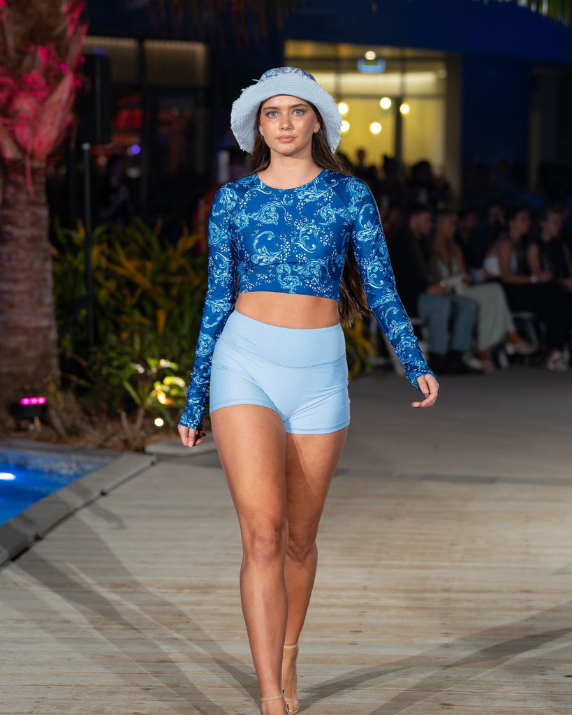 women on catwalk wearing white and blue crop rash guard and light blue swim shorts with bucket hat