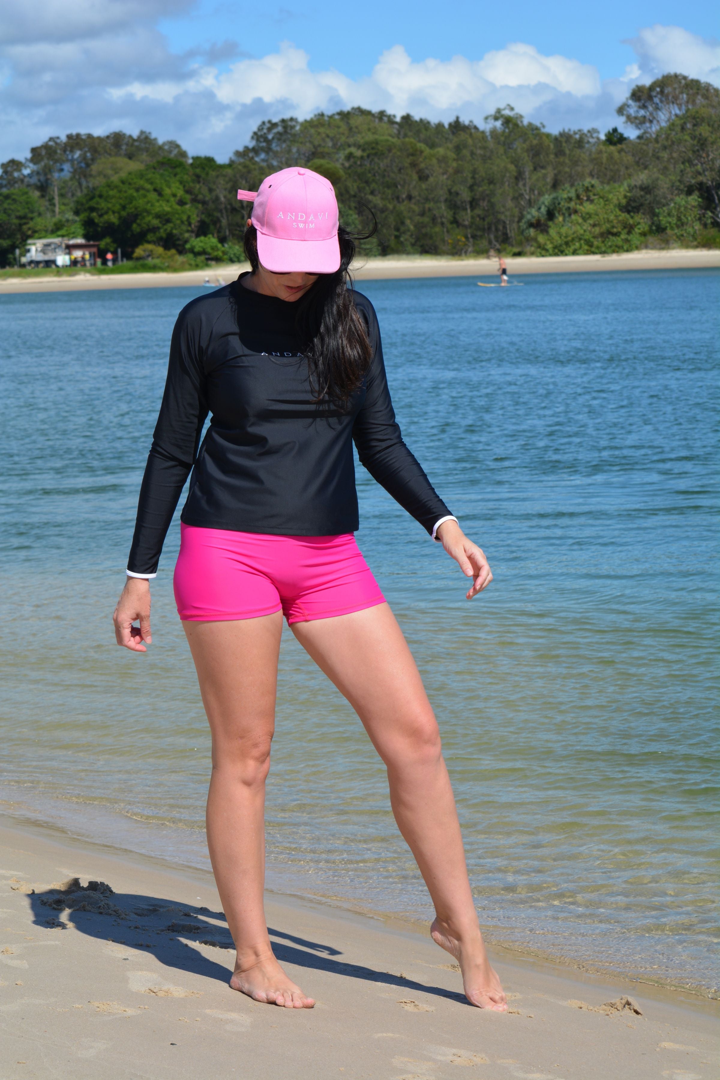 Person wearing a black long-sleeve shirt and pink shorts standing on a beach with water and trees in the background.