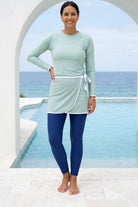 Woman wearing a light green long-sleeve top and blue leggings standing by a pool with ocean view.