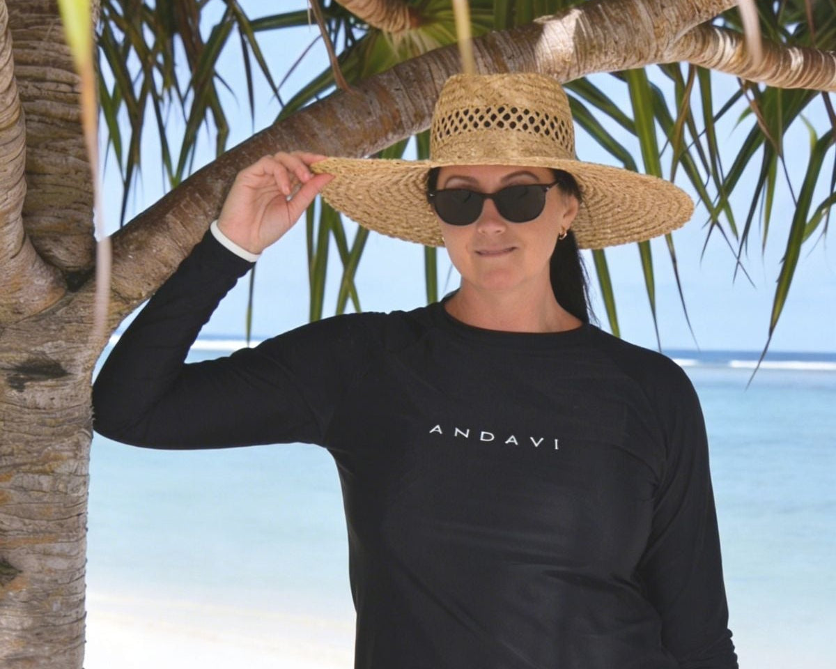 Woman wearing a black long-sleeve rash guard and shorts on a beach with palm trees.
