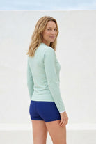 back view of women standing in front of white wall wearing green andavi rash guard and blue shorts