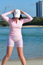 women with her hand on her head on the beach wearing long sleeve light pink rash guard and floral bike shorts 