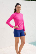 Woman wearing a pink long-sleeve top and blue shorts standing against a white wall.