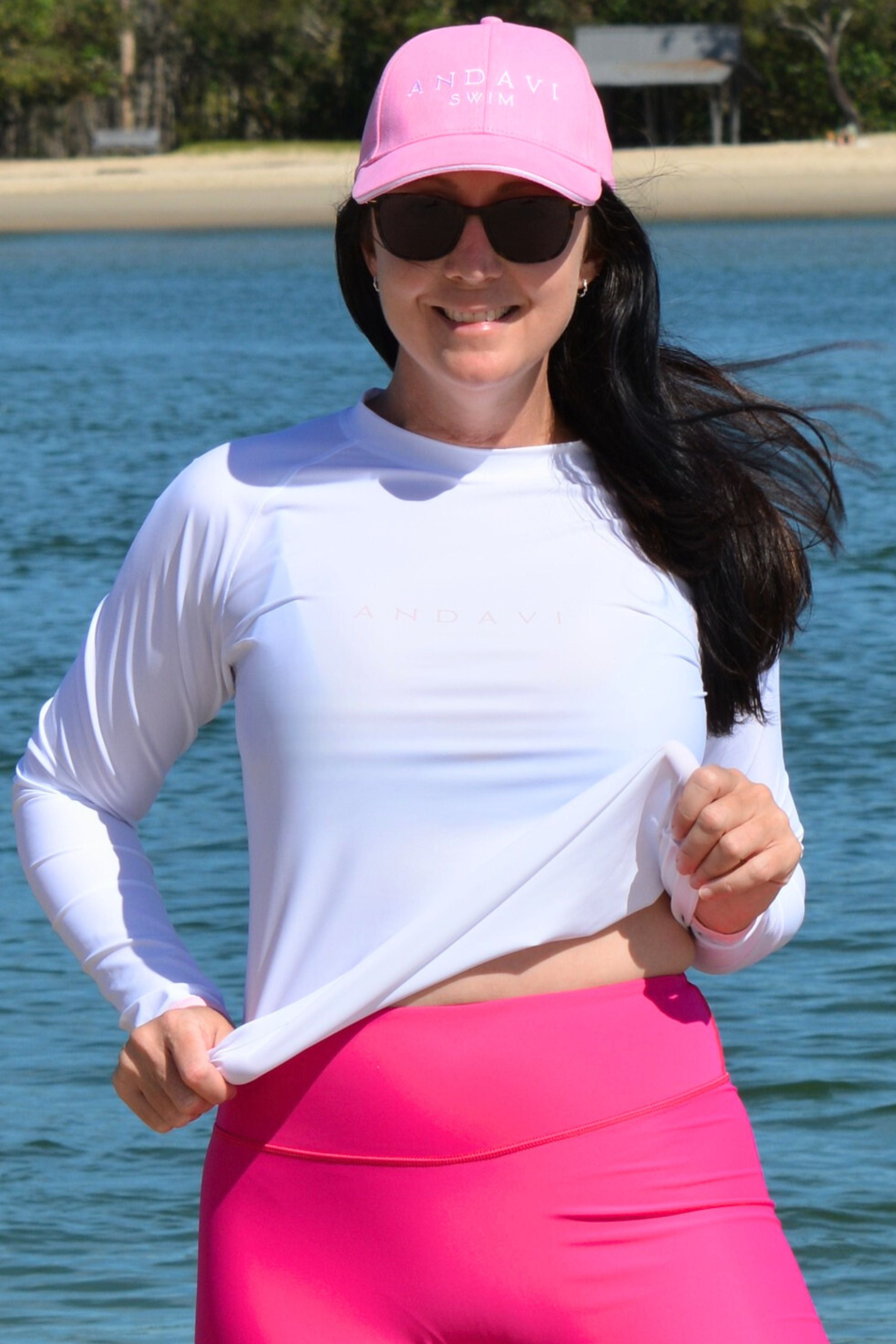 Woman wearing a white long-sleeve top and pink pants with a pink cap and sunglasses, standing by a body of water.