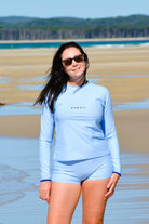 Woman wearing a light blue swimsuit with 'ANDAVI' branding on a beach.