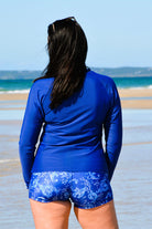 back view of women on beach wearing dark blue long sleeve rash guard and blue and white print swim shorts