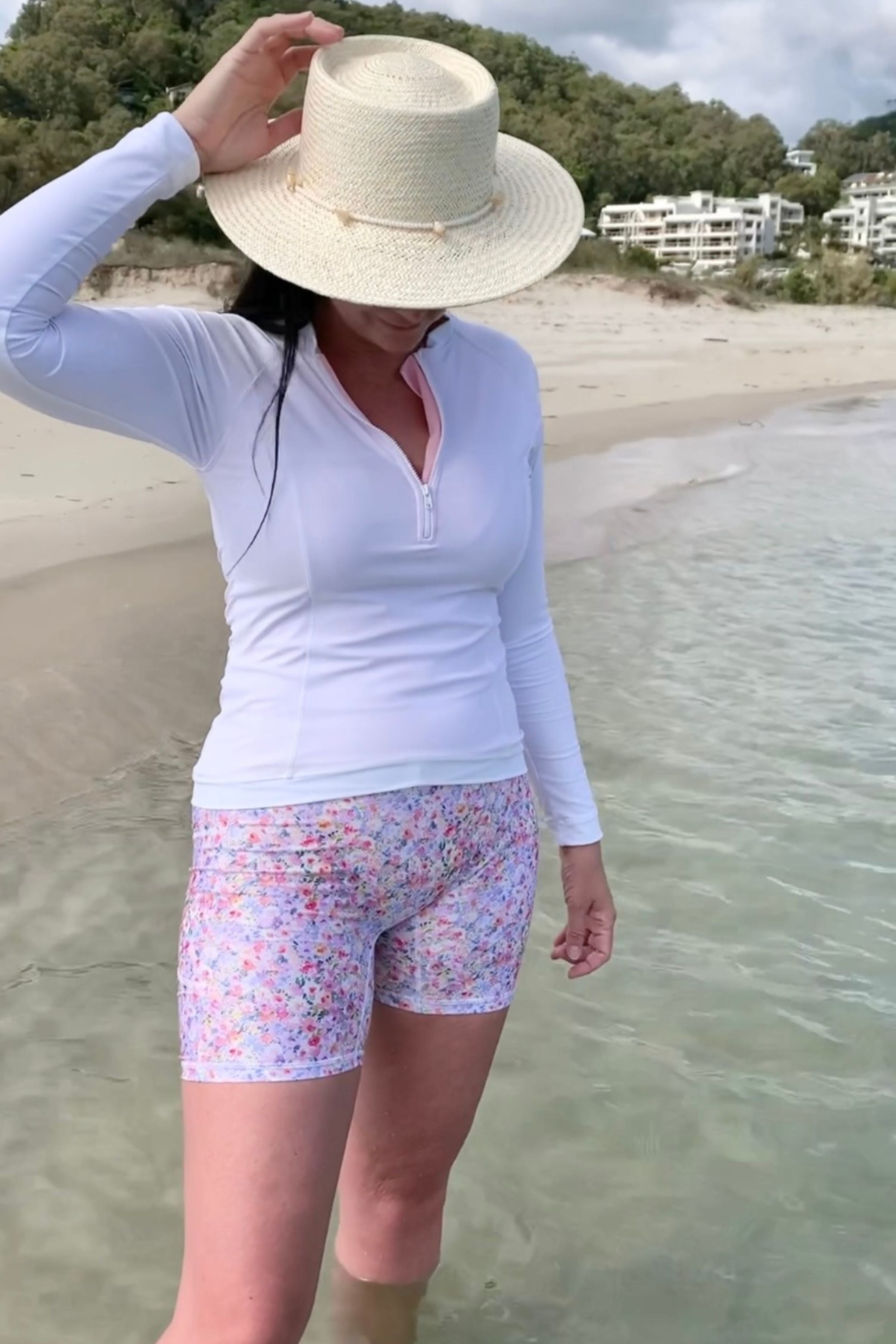 Person wearing a white long-sleeve shirt, floral shorts, and a straw hat on a beach.